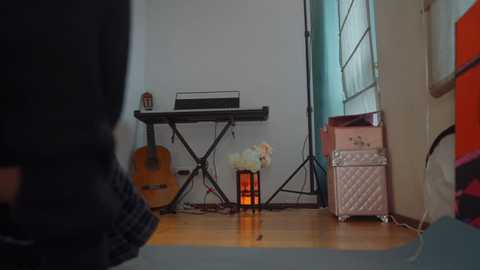 Video of a cozy, cluttered room with a black keyboard, a guitar, and a pink suitcase. A person's legs are visible in the foreground, wearing dark pants. The walls are light-colored, and a window with light curtains lets in natural light.