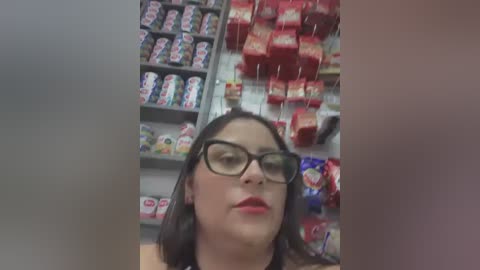 Media: A video shows a woman with fair skin and shoulder-length dark hair, wearing glasses and red lipstick, looking downward in a grocery aisle filled with red and white candy packets and snack bags.