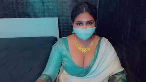 Media: Video of a South Asian woman with dark hair, wearing a teal salwar kameez with a plunging neckline, white dupatta, and a yellow floral necklace, sitting on a bed with dark bedding, against a textured black wall.