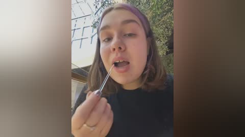 Media: A video of a young woman with light skin and shoulder-length brown hair, wearing a black sweater, holding a fork in her mouth, standing outside next to a white building and green foliage.