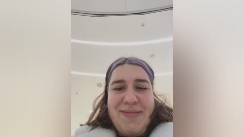 Media: Video of a young woman with light skin and shoulder-length brown hair, wearing a purple headband and a white fur-trimmed jacket, smiling slightly. She is inside a sleek, white, curved elevator with round ceiling lights and mirrored walls.