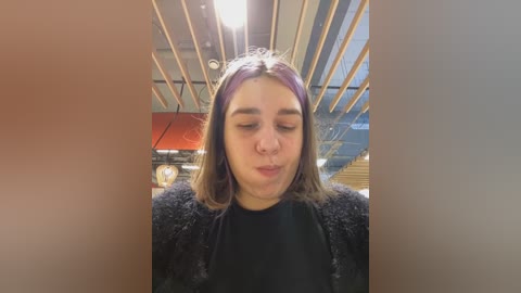 Media: Video of a young woman with straight, shoulder-length hair, wearing a black top and a fuzzy black cardigan, standing indoors under a wooden ceiling with exposed beams.