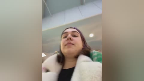 Media: A video of a young woman with medium skin tone, brown hair, and a black top, wearing a white fur-lined jacket. The background shows an indoor space with a green chair and a white ceiling.