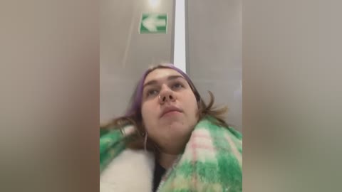 Media: Video of a young woman with light skin, wearing a green and white plaid blanket and a purple headband, looking slightly upward with a neutral expression. Background shows a white door and a green exit sign.