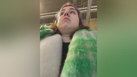 Media: A video of a young woman with fair skin, light brown hair, and a pink headband, wearing a green and white fuzzy blanket. She looks upward, with a neutral expression. The background features industrial pipes.