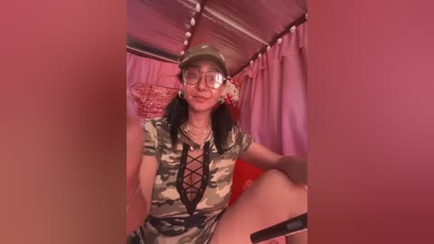 Media: Video of a woman in a camouflage dress with lace-up front, wearing a green cap, glasses, and sitting on a bed with pink curtains, smiling.