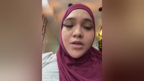 A video of a young woman with medium skin tone, wearing a maroon hijab and gray shirt, eyes closed in a contemplative expression, in a softly lit indoor setting.