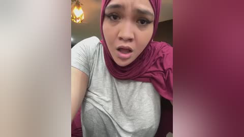 Video of an Asian woman with medium skin tone, wearing a grey t-shirt, a burgundy hijab, and a burgundy scarf, with a surprised expression. Background shows a dimly lit room with a pendant light.