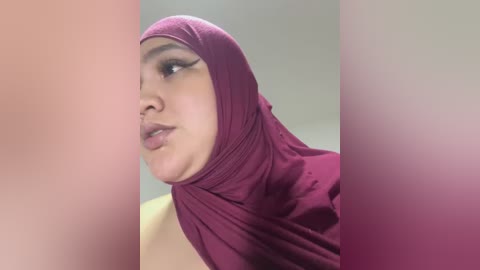 Media: Video of a young woman with light brown skin and full lips, wearing a maroon hijab and looking to the side. Background is a blurred gradient of pink and white.