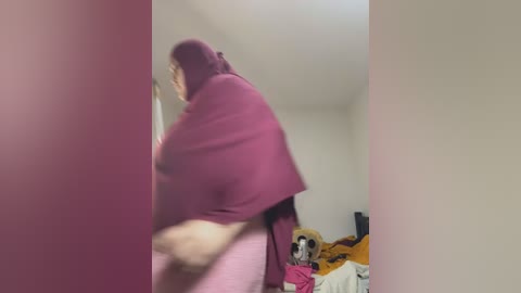 Media: A blurry video shows a woman in a maroon headscarf and pink dress walking away from the camera, with a cluttered, off-white room in the background.