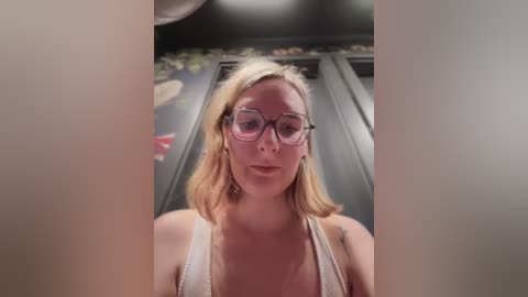 Video of a Caucasian woman with light blonde hair, wearing glasses and a white tank top, standing in a dimly lit room with floral wallpaper.