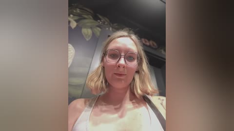 Media: Video of a Caucasian woman with shoulder-length blonde hair, wearing glasses, a white halter top, and a beige shoulder bag. She stands indoors, with a blurred background featuring decorative leaves and a circular design.