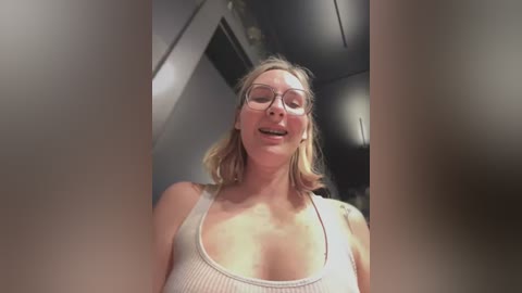 Video of a smiling, fair-skinned blonde woman with shoulder-length hair, wearing clear glasses and a white tank top, taken from a low angle. The background features modern, industrial elements.