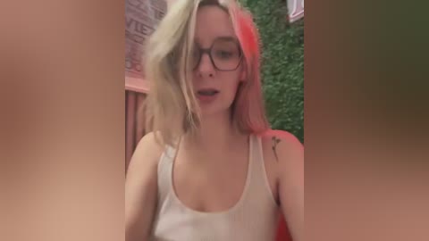 Media: Video of a young Caucasian woman with shoulder-length, wavy, blonde hair, wearing black-rimmed glasses and a white tank top. She has a small tattoo on her left shoulder. The background features a green wall and a blurred, partially visible person.