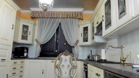 Media: A video of a stylish kitchen with white cabinets, a tufted chair, a window adorned with sheer gray curtains, and a metallic faucet.