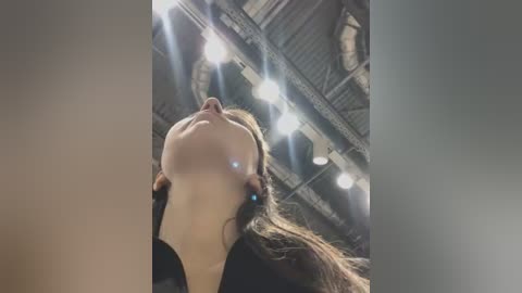 Media: Video of a woman with long brown hair, looking up, captured from a low angle, with studio lights illuminating her face and ceiling.