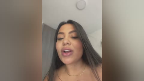 Media: Video of a Latina woman with long, straight black hair, light brown skin, and full lips. She has makeup with dark eyeliner and wears a beige top. The background is blurred, showing a white ceiling and a soft, muted color palette.