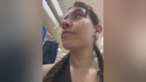 Media: Video of a young woman with light skin, short brown hair, and a black top, looking upward with a slight smile. Background includes a blue curtain and fluorescent lighting in a dimly lit room.