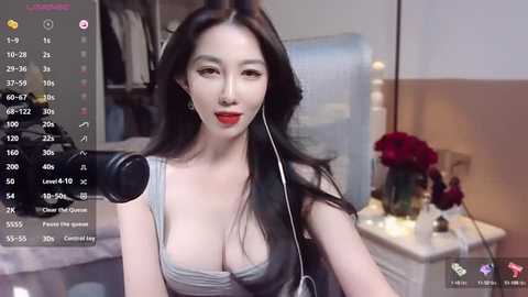 Media: Video of an Asian woman with long black hair, wearing a low-cut gray top, recording herself on a webcam. Background shows a bedroom with a bed, flowers, and a nightstand.