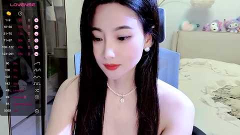 Media: Video of a young Asian woman with long black hair, fair skin, and red lipstick, sitting in a bedroom with a bed, stuffed animals, and a blue chair.