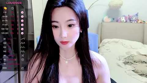 Media: A video of an East Asian woman with long, straight black hair, wearing a pearl necklace, and a black top, in a bedroom with a bed and stuffed toys.