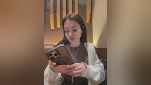 Media: Video of an Asian woman with long black hair, wearing a beige jacket and brown top, taking a selfie with her iPhone, in a dimly lit, modern interior with beige walls and vertical wooden panels.