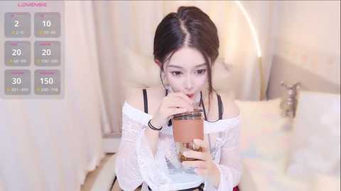 Media: Video of an Asian woman with long dark hair, wearing a white lace top, drinking from a cup, in a soft-lit bedroom.