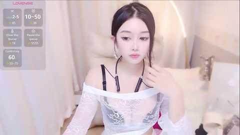 Media: A video of an East Asian woman with fair skin and long, straight black hair, wearing white lace lingerie, sitting on a bed in a softly lit bedroom.