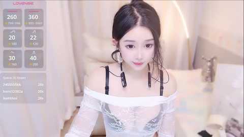 Media: Video of an East Asian woman with fair skin and long black hair in a white lace off-shoulder top, wearing black tassel earrings, in a softly lit bedroom.