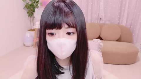 Media: A video of an East Asian woman with straight, shoulder-length black hair and pale skin, wearing a white face mask and a white sleeveless top, in a pink-lit, minimalist room with beige couches.