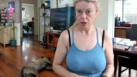 Media: Video of a middle-aged, fair-skinned woman with blonde hair, wearing a blue tank top, seated indoors with a dog beside her. Background includes a TV, shelves, and a kitchen area.