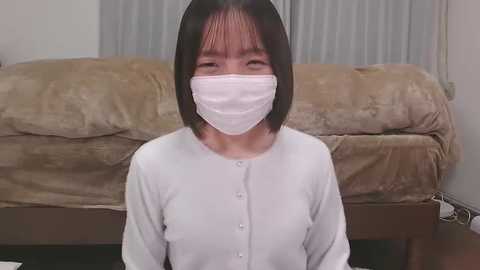 Media: A video of a young Asian woman with short black hair, wearing a white mask, white long-sleeve top, and beige blanket, standing indoors against a beige sofa and white curtain background.