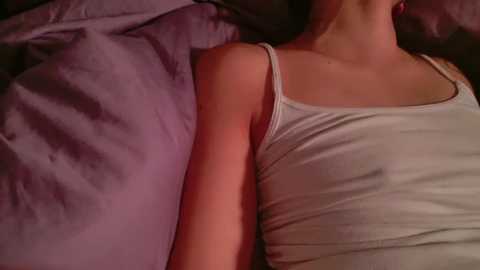 Media: A video of a woman lying on her side, wearing a white tank top, her bare shoulder exposed, with a purple pillow behind her.