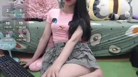 Media: A video shows an Asian woman with long black hair, wearing a pink T-shirt and a black and white checkered skirt, sitting on a green rug in a room with plush toys and a digital clock.