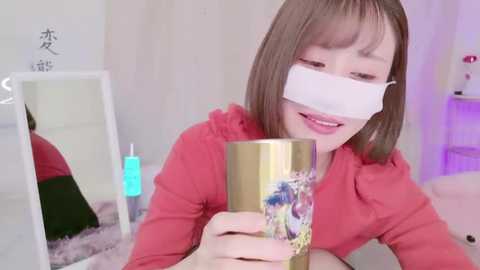 Media: A video of an Asian woman with a face mask, wearing a red hoodie, drinking from a gold cup with floral design, in a cozy, softly lit room with a mirror, plush toys, and Japanese text.