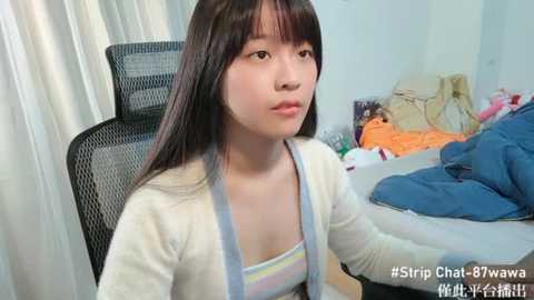 Media: Video of a young Asian woman with long black hair, wearing a white cardigan over a striped tank top, sitting on a black mesh chair in a messy bedroom with clothes and a blue blanket in the background.