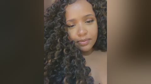 Media: Video of a young Black woman with medium brown skin, sporting a voluminous, curly afro hairstyle. She has a serene expression, with subtle makeup. The background is blurred, focusing attention on her.