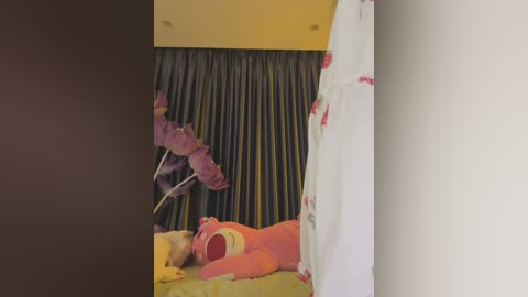 Media: Video of a child lying on a plush, colorful bedspread with a teddy bear and purple flowers in the background, partially hidden by a white curtain.