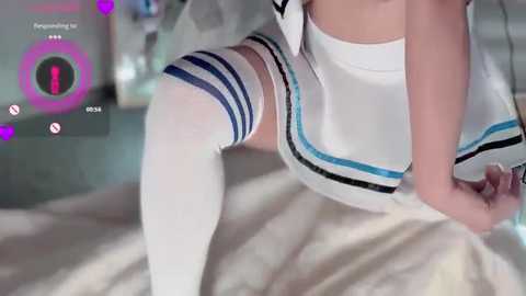 Media: Video of a woman in a white, high-waisted skirt with blue stripes, wearing thigh-high white socks, sitting on a bed with a beige sheet. The background shows a virtual reality interface with \"Eroge\" and \"10:02\" displayed.