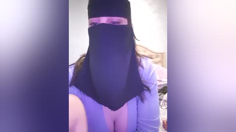 Media: A video of a woman with long brown hair, wearing a black face veil, light blue shirt, and a black headscarf. The background shows a bedroom with a bed and pillows.