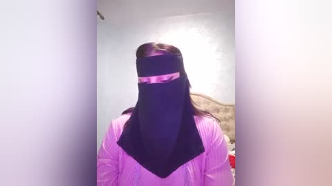 Media: A video of a person wearing a black hijab and a light pink shirt, standing in a dimly lit room with white walls and a beige headboard visible in the background.