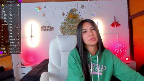 Media: Video of a young woman with long black hair, wearing a green hoodie, sitting on a white chair in a brightly lit room with Christmas decorations.