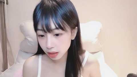 Media: Video of a young Asian woman with straight black hair and fair skin, wearing a white spaghetti-strap top, seated in a beige room with a white pillow and wooden door in the background.