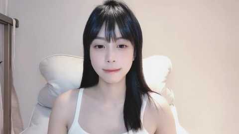 Media: Video of an East Asian woman with long black hair, wearing a white tank top, sitting on a bed with white pillows. The background is a plain, light-colored wall.