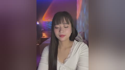 Media: Video of a young East Asian woman with long black hair, wearing a white lace-trimmed top, seated in a dimly lit room with purple and orange lighting.