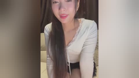 Media: Video of an Asian woman with long black hair, wearing a white lace-trimmed top, and black shorts, sitting on a bed with beige sheets, in a dimly lit room with dark curtains.