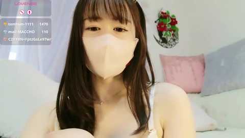 Media: Video of an Asian woman with long dark hair and pale skin, wearing a surgical mask, pink bra, and sitting on a bed with white and pink pillows.