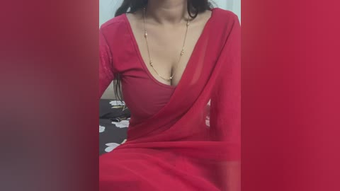 Media: A video of a woman in a red saree with a deep neckline, revealing ample cleavage. She wears a gold necklace and sits on a bed with a patterned blanket.