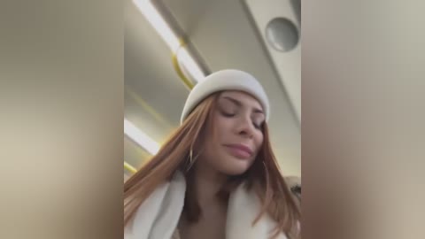 Media: Video of a young woman with light skin, long brown hair, and a white beanie, wearing a white coat, standing in an airport, with blurred background and fluorescent lights.