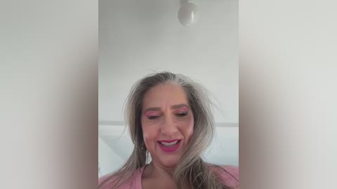 Media: A video of a middle-aged woman with shoulder-length, wavy gray hair, wearing pink eyeshadow and lipstick, smiling with closed eyes, against a white background with a hanging light bulb.